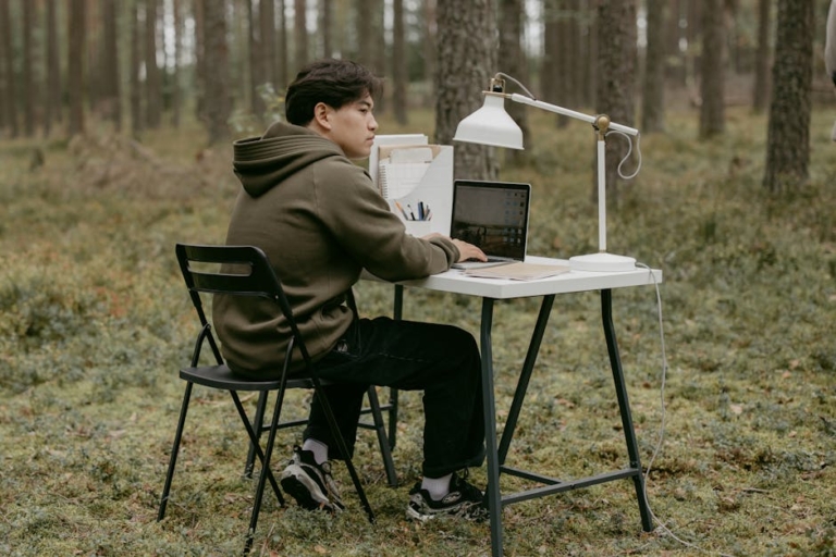 Man working on laptop at an outdoor desk in a forest setting, embracing nature.